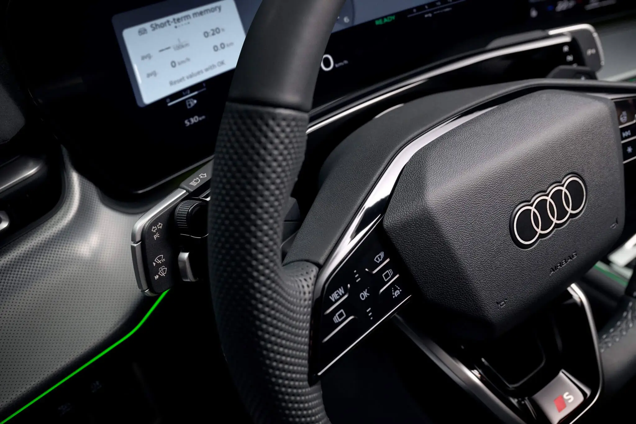 audi q3 interior volante Detalhe do volante do Audi Q3 2026 — logótipo Audi, comandos multifunções, painel de instrumentos digital e iluminação ambiente verde.