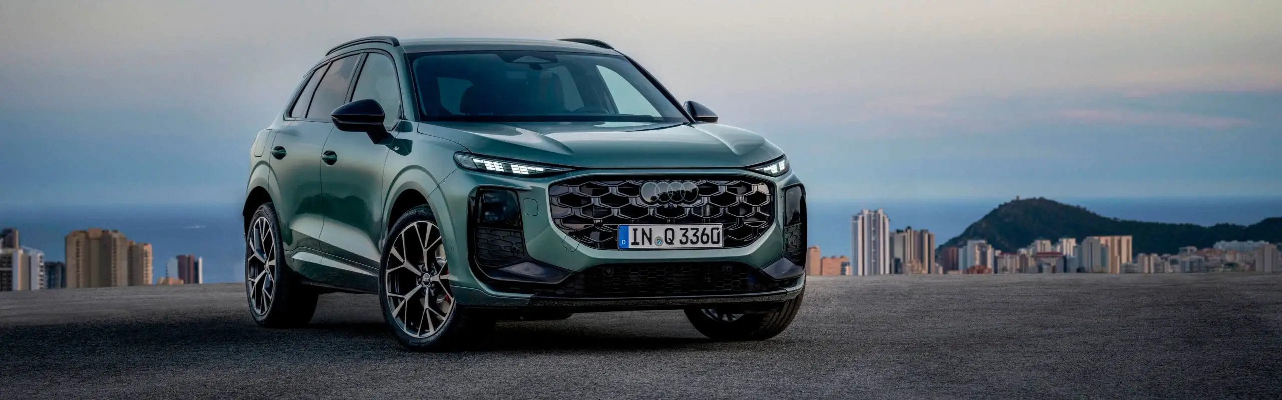 Audi Q3 2026 verde, vista frontal em três-quartos, SUV estacionado com cidade litoral e mar ao fundo.