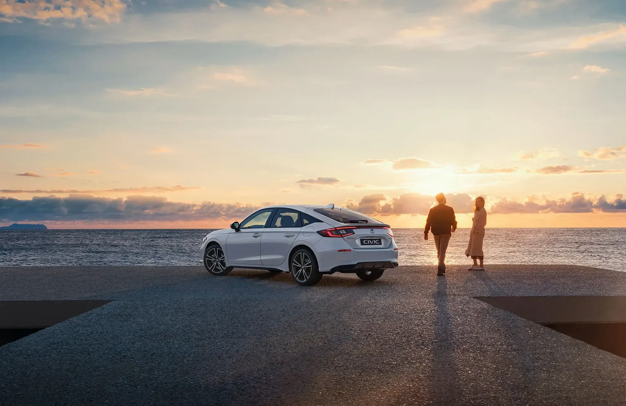 Casal junto a um Honda Civic branco numa paisagem de pôr do sol à beira mar